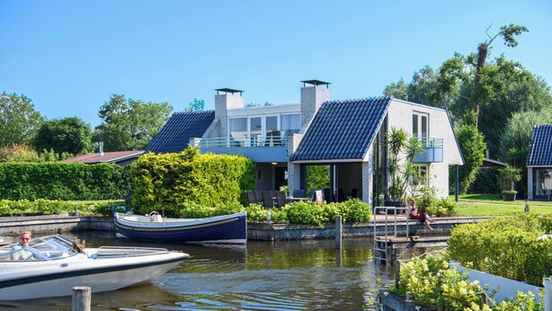 A houseboat docked at a canal with a modern design, surrounded by lush greenery and a clear blue sky.