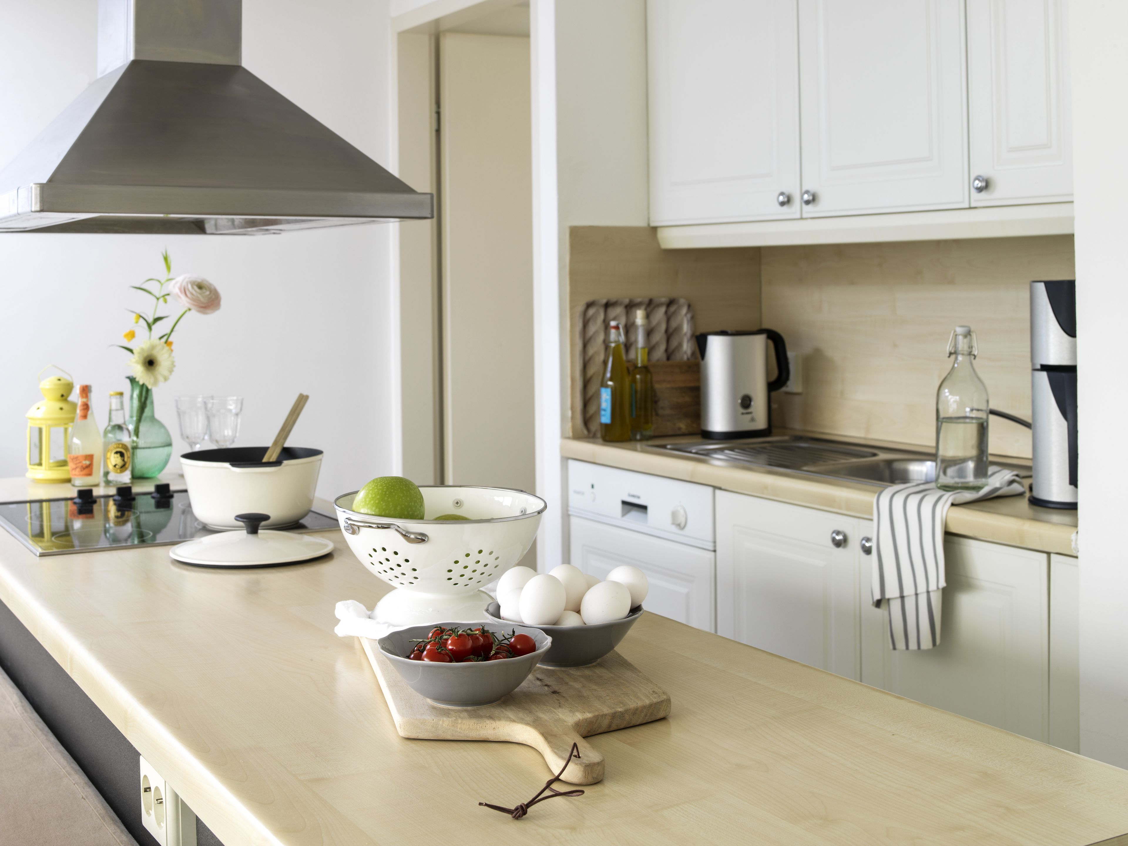 A kitchen with white cabinets, a stainless steel range hood, a sink, and various kitchen items like a colander, a bowl of eggs, and a cutting board with a knife.