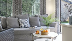 A modern living room with a grey sofa, patterned cushions, a small round table with a plant and fruit basket, and a view of trees through patterned curtains.