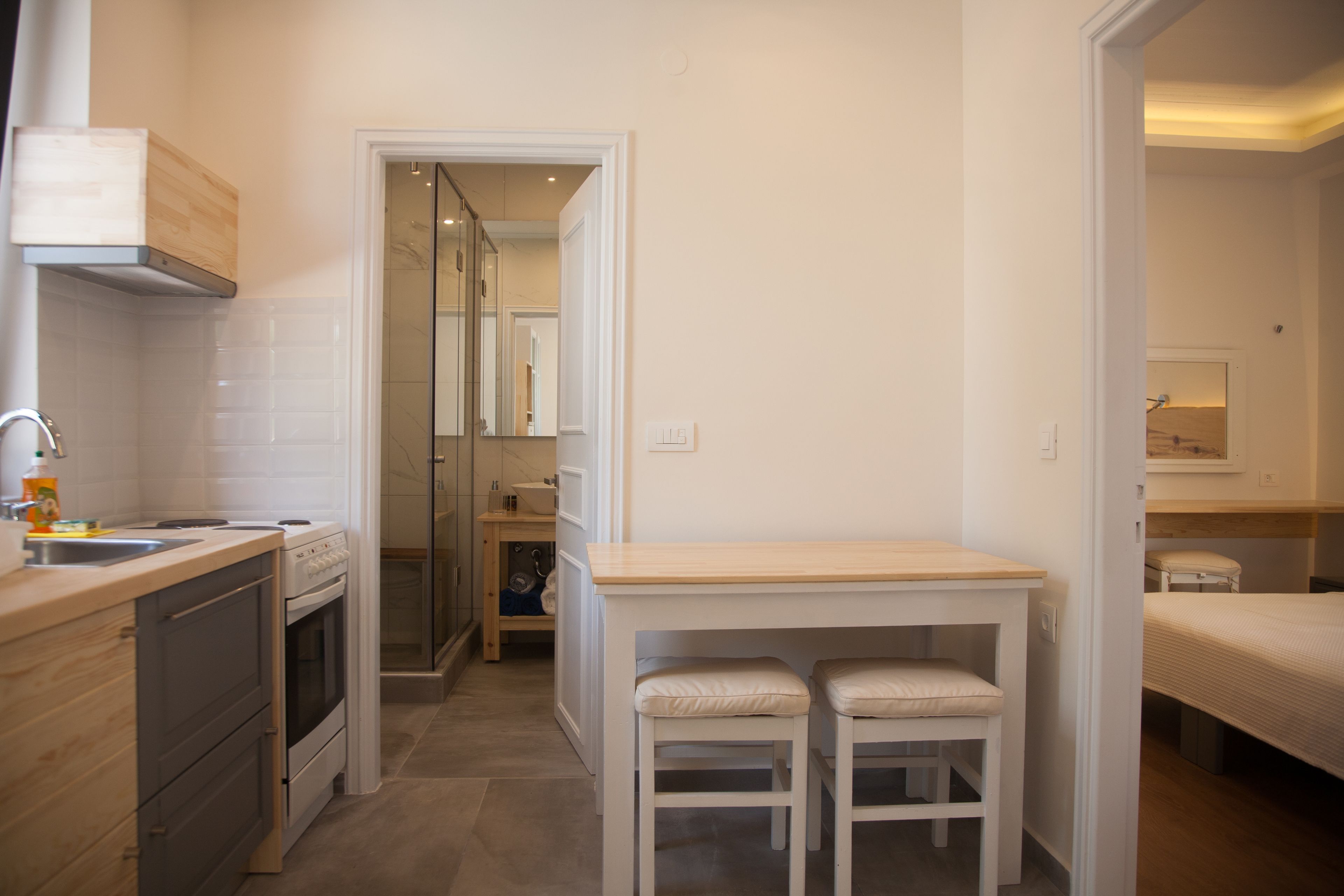 A compact kitchen with a stove, sink, and wooden countertop. There is a small dining area with a table and two chairs. A hallway leads to a bedroom with a bed and a nightstand.