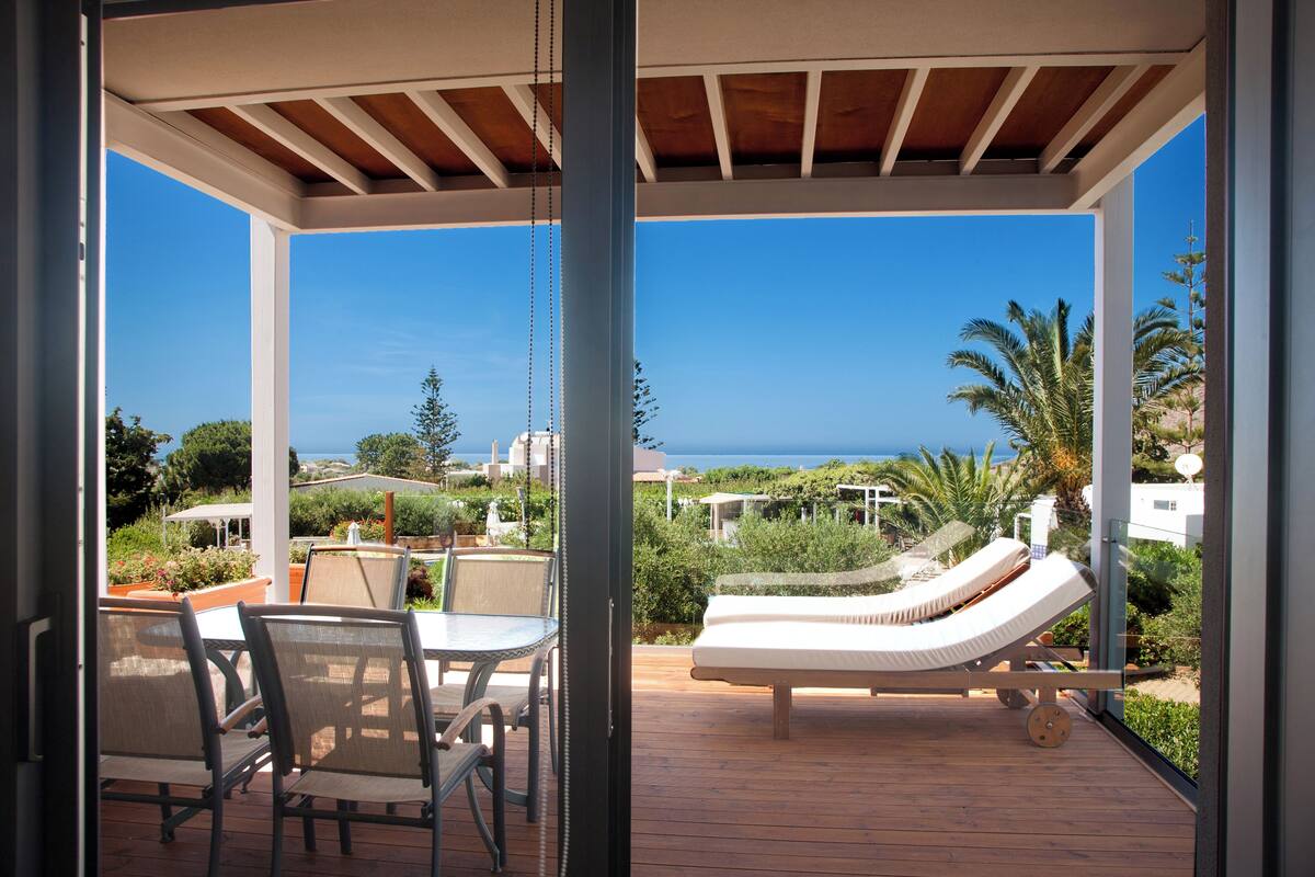 A covered outdoor area with a table and chairs, a lounge chair, and a view of the ocean.