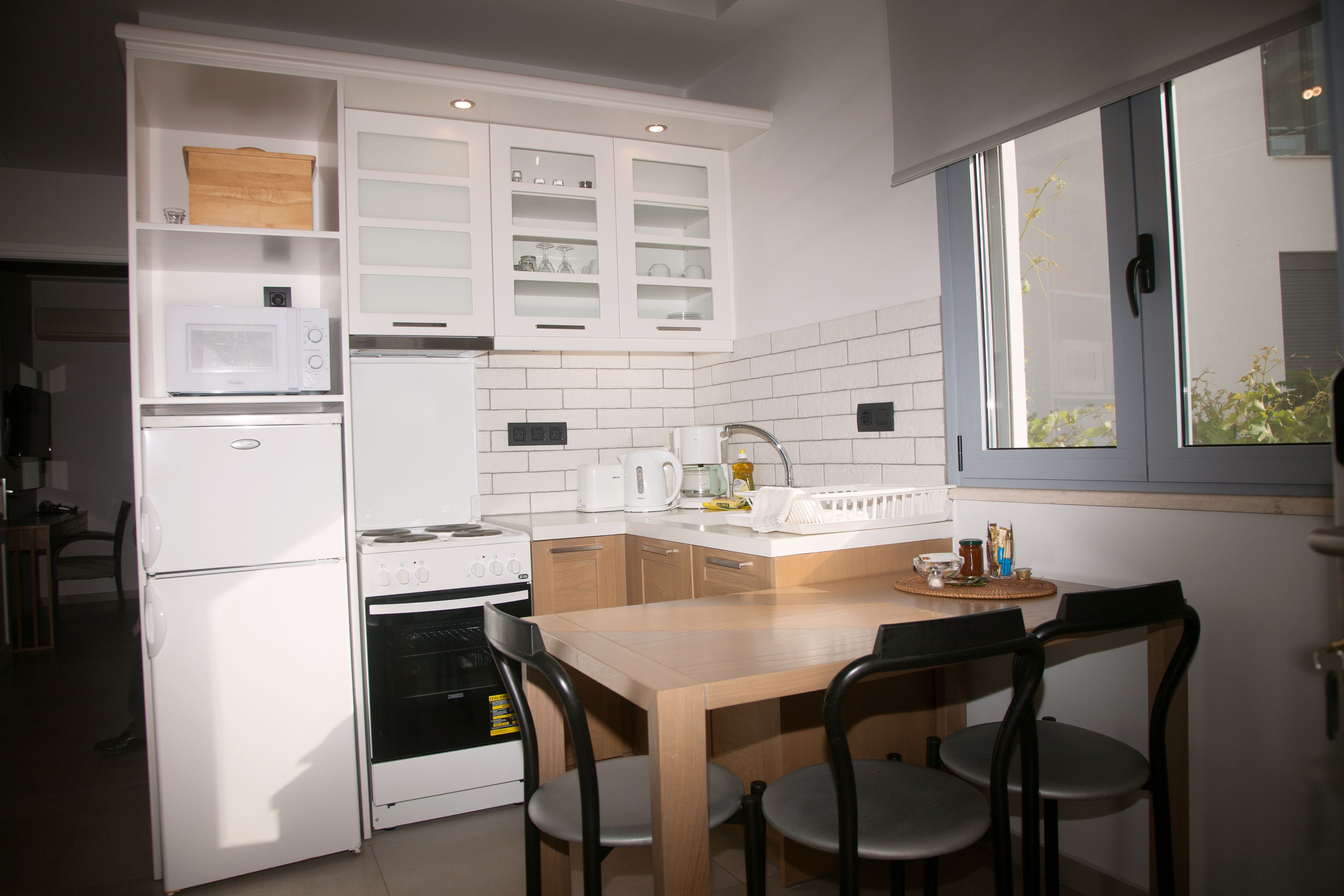 A compact kitchen with white cabinetry, a microwave, a stove, and a sink. There is a dining table with chairs and a window providing natural light.