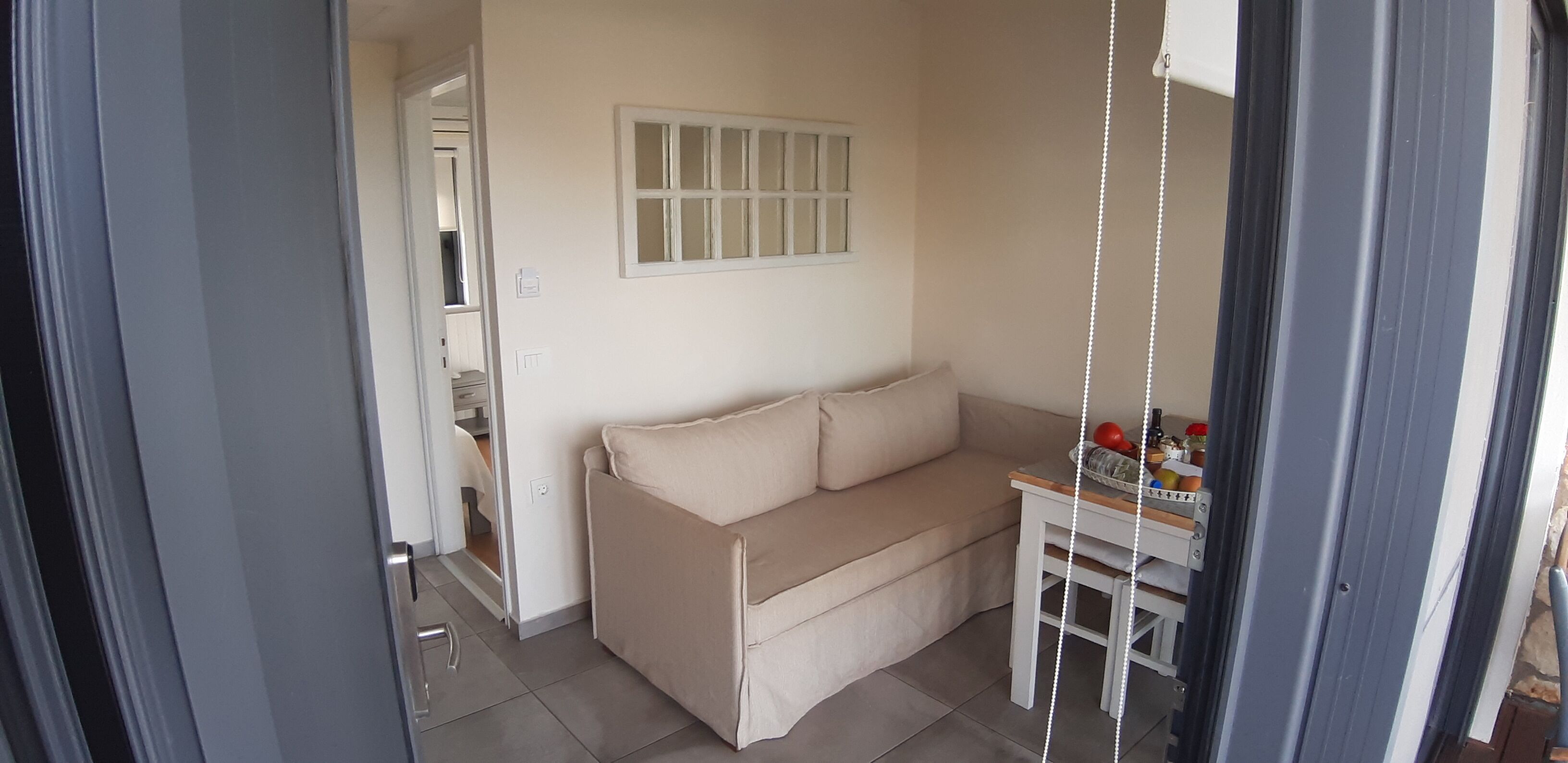 A small, cozy room with a beige sofa, a small table with a basket, and a window with blinds.