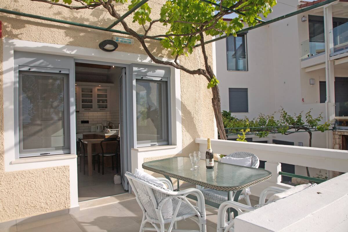 A balcony with a glass table and white wicker chairs, overlooking a building with a kitchen and dining area.