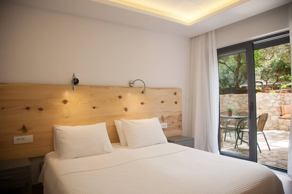 A hotel room with a wooden headboard, a bed with white bedding, a nightstand, and a view of an outdoor patio with a table and chairs.