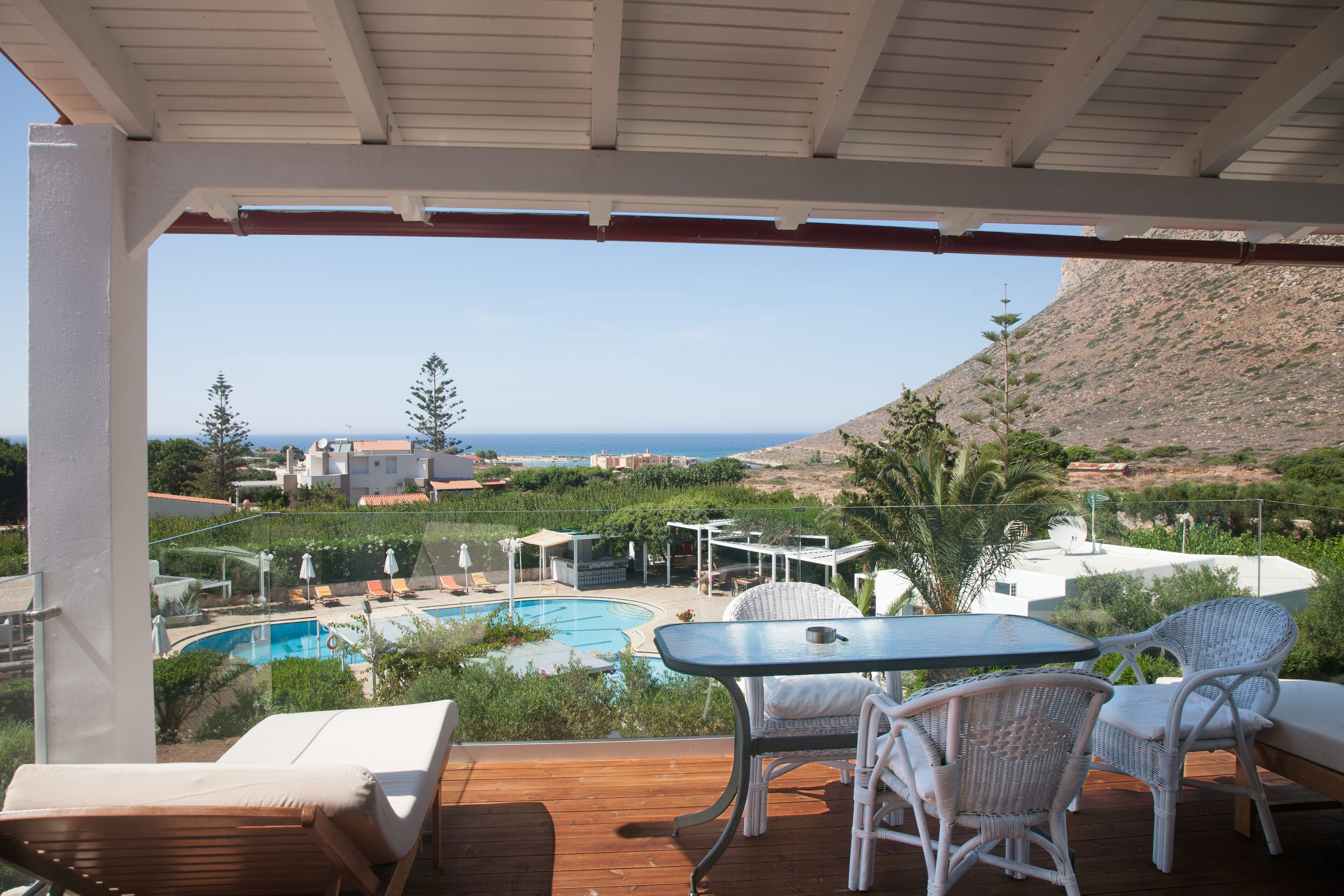 A wooden deck with a glass table and white wicker chairs, overlooking a pool and a mountainous landscape.