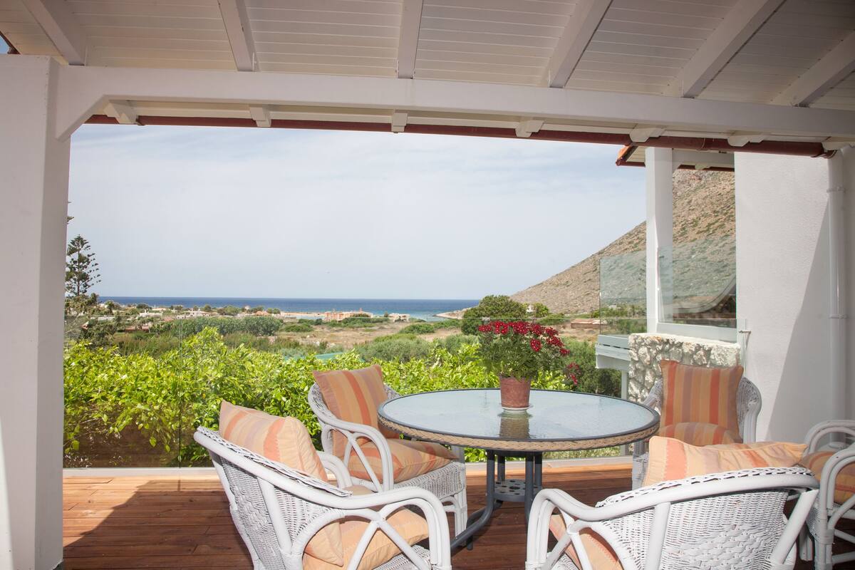 A covered patio with wicker furniture and a glass table, overlooking a scenic landscape with hills and a view of the sea.