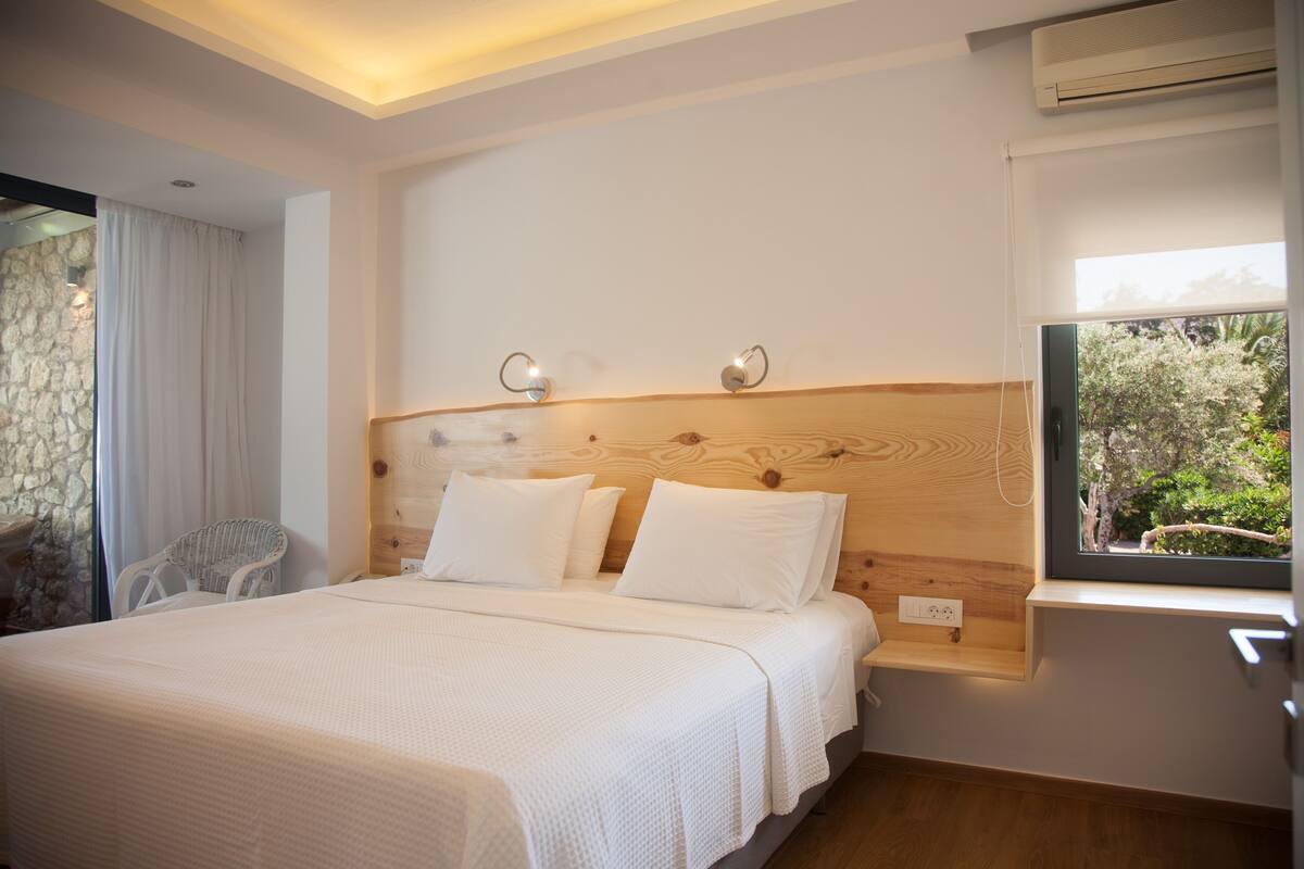 A bedroom with a wooden headboard, a large bed with white bedding, a wicker chair, and a window with a view of greenery.