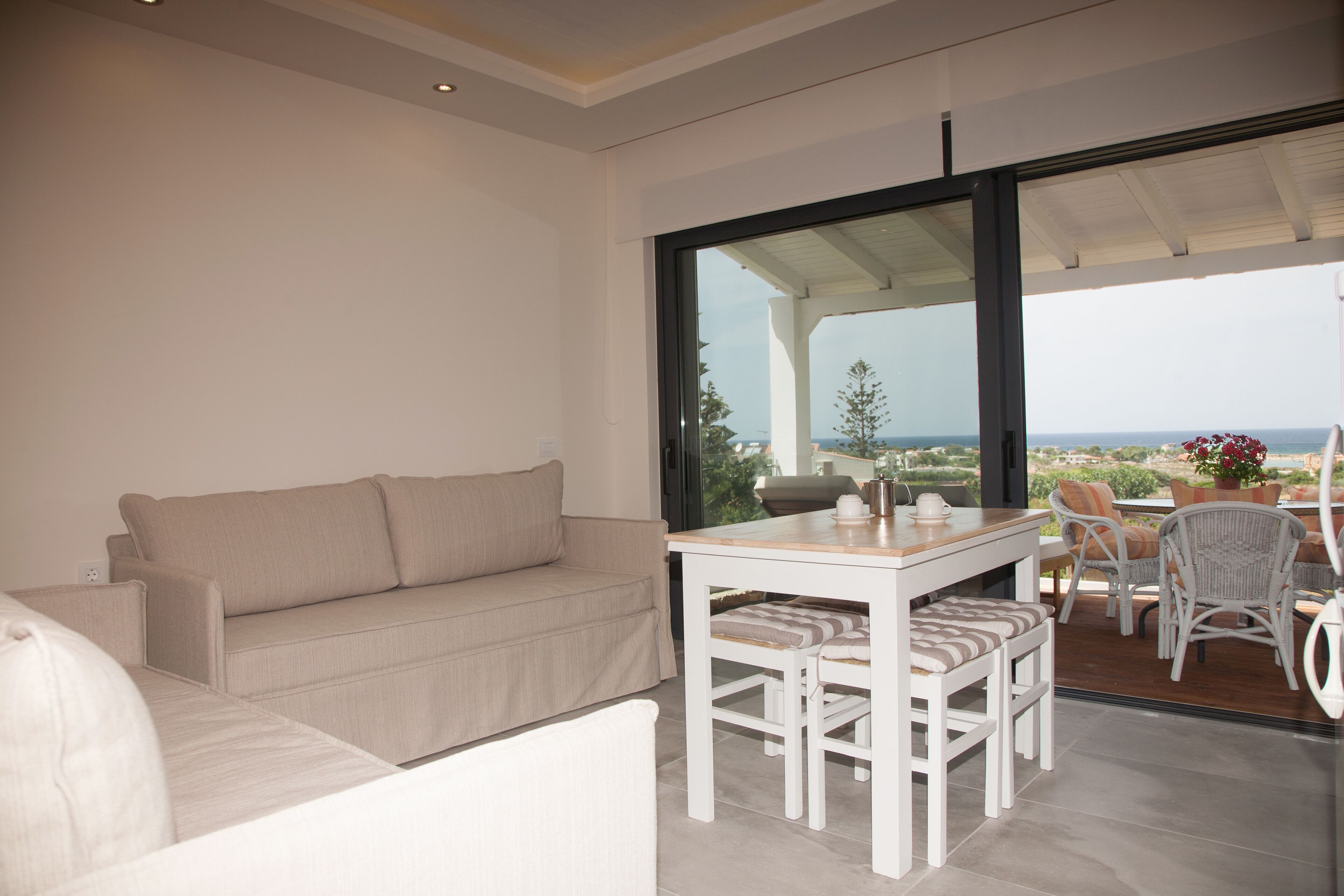 A modern living room with a beige sofa, a white dining table with chairs, and a large glass door leading to an outdoor patio with wicker furniture and a view of the landscape.