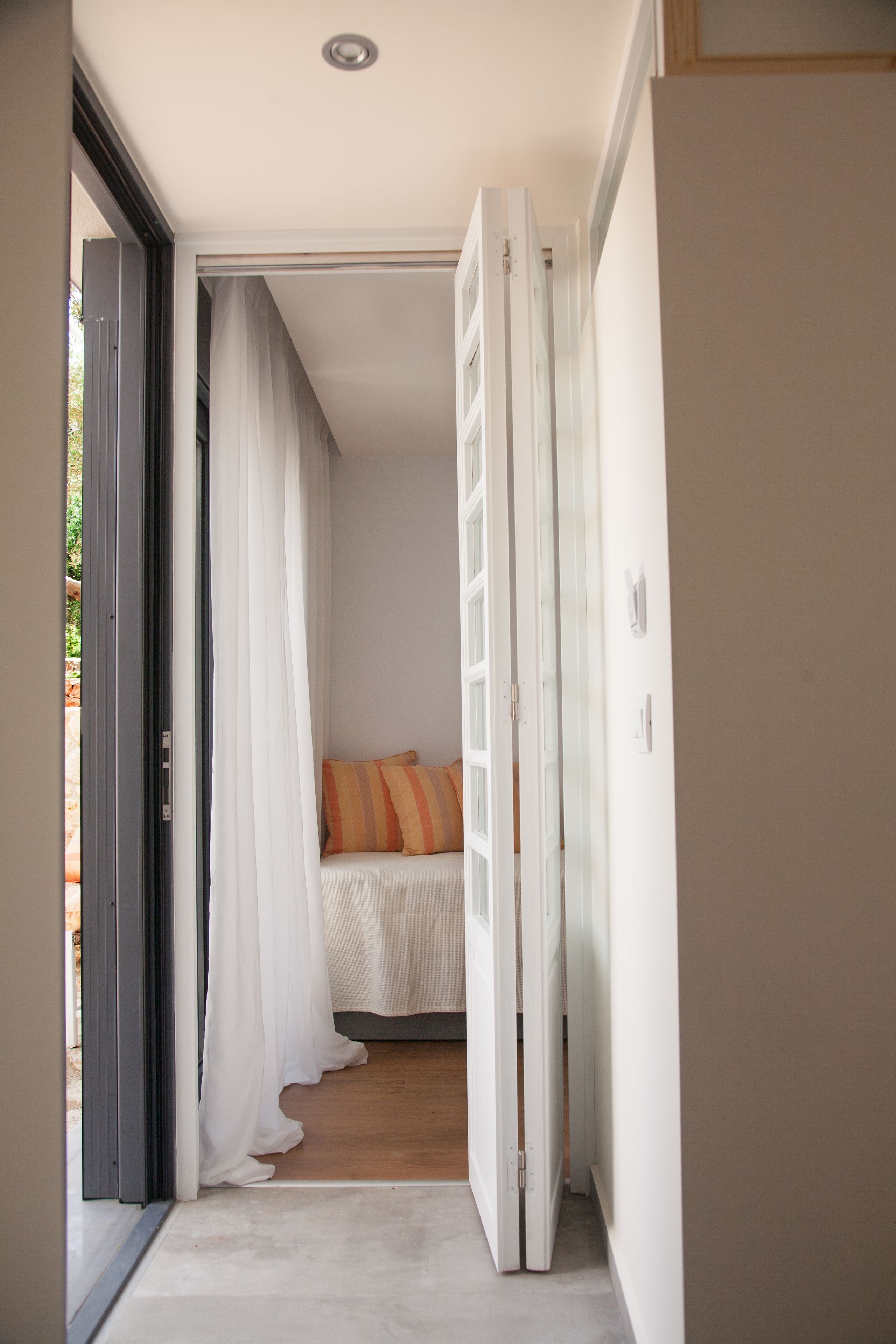 Un couloir étroit mène à une chambre à coucher qui contient un lit, des rideaux blancs et un plancher en bois.