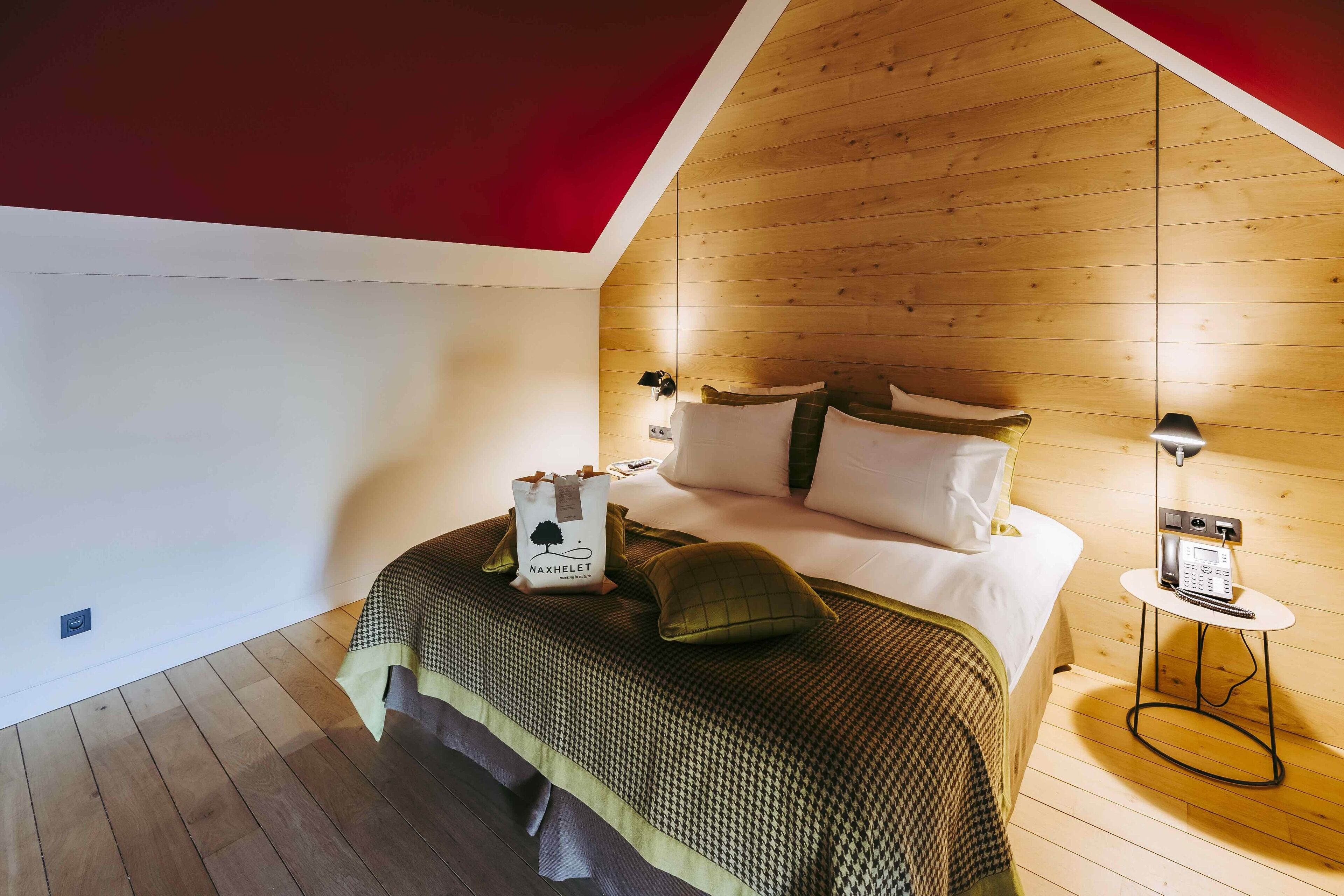 A bedroom with a wooden ceiling, a bed with a patterned bedspread, pillows, a bedside table with a lamp, and a bag on the bed.