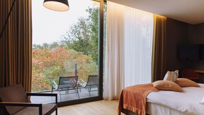 Quarto de hotel com cama, cadeira, mesa e uma grande janela que oferece vista para árvores e um espaço de terraço.