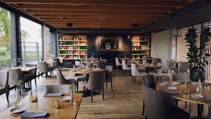 Restaurante com mesas e cadeiras de madeira, janelas amplas e prateleiras com garrafas.