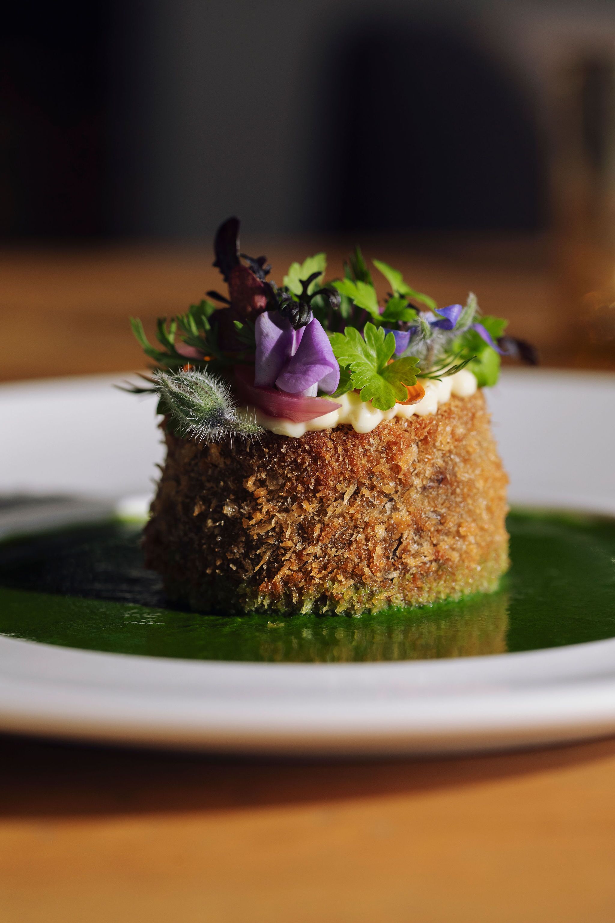 A dish with a crispy base, topped with greens and purple flowers, served on a white plate with a green sauce.