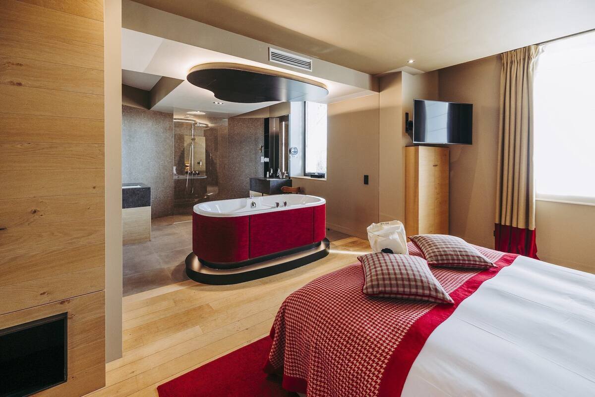 A modern hotel room with a large bed, a red and white checkered bedspread, a red velvet bath tub, a large mirror, and a flat-screen TV mounted on the wall.