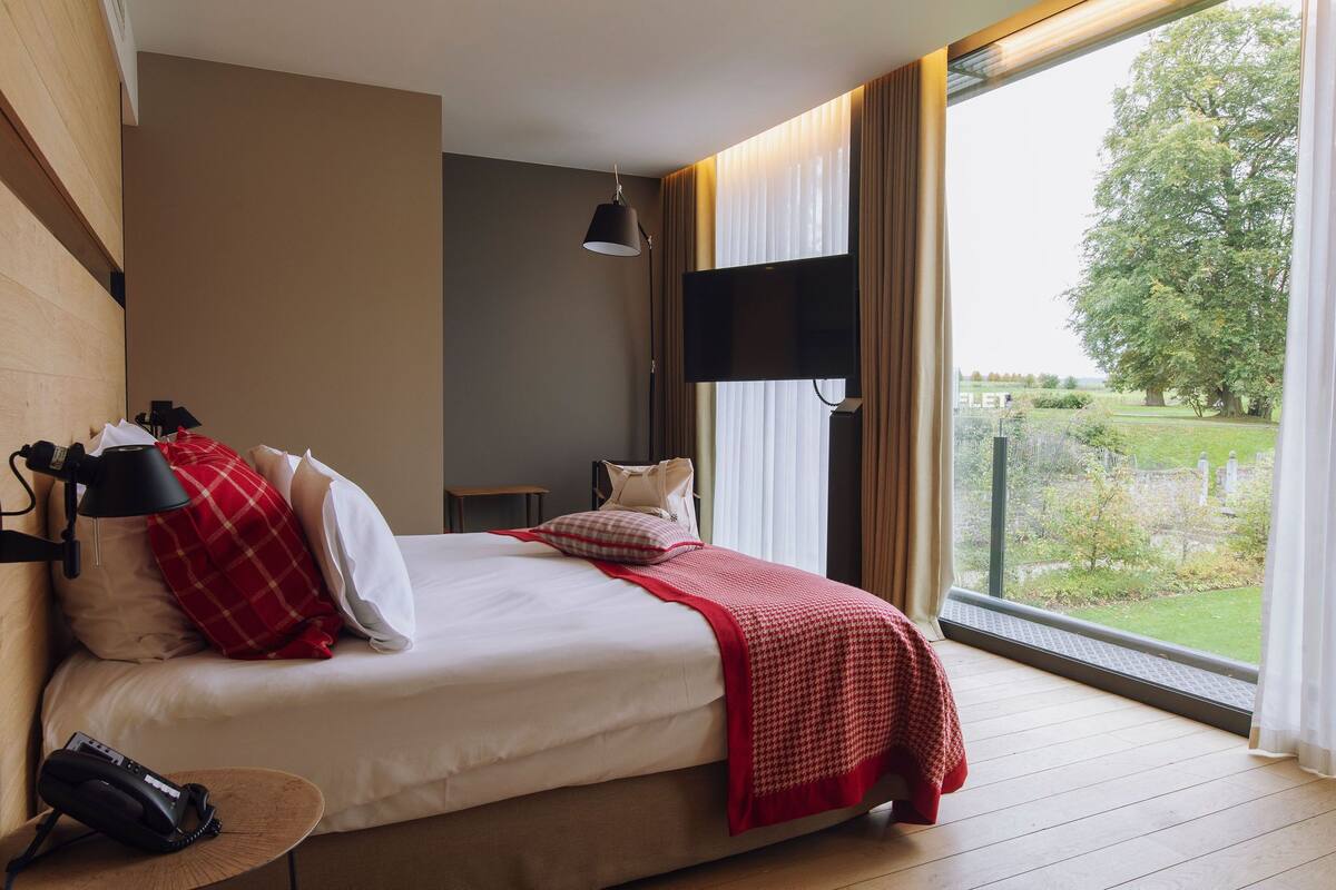 A bedroom with a bed, a bedside table with a lamp, a telephone, and a window with a view of greenery.