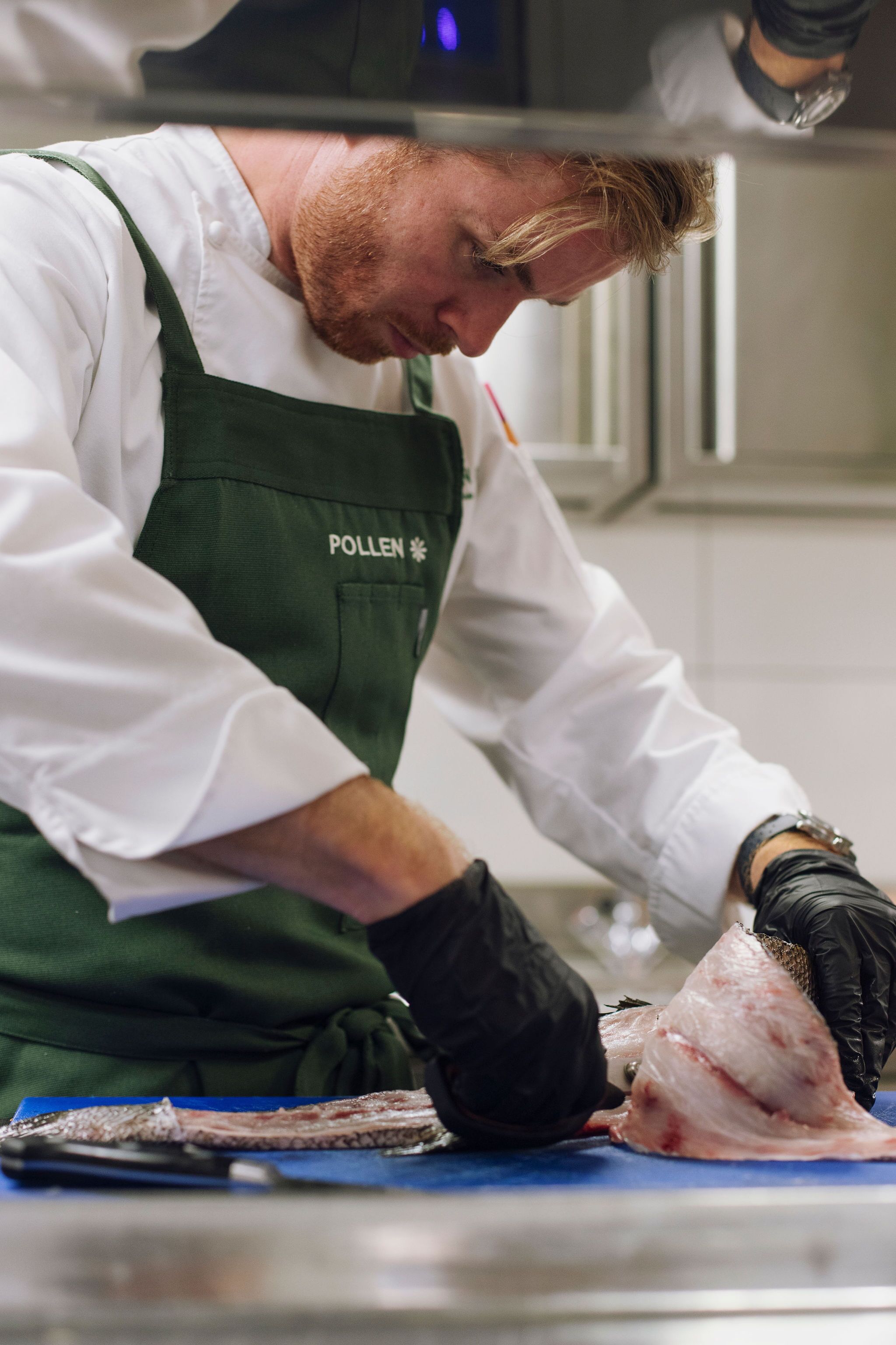 En person i en vit skjorta och ett grönt förkläde som arbetar med fisk på en blå skärbräda i ett kök.