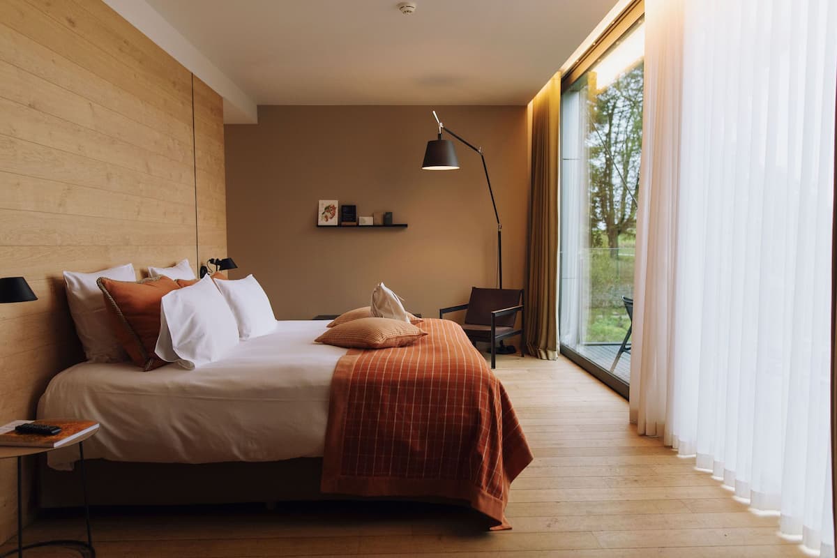 A modern hotel room with a large bed, a desk with a lamp, a chair, and a view of greenery through large windows.