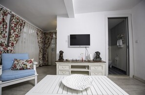 Una habitación con un sofá blanco, una mesa de centro blanca, un televisor en la pared, un aparador de madera y un baño visible a través de una puerta abierta.