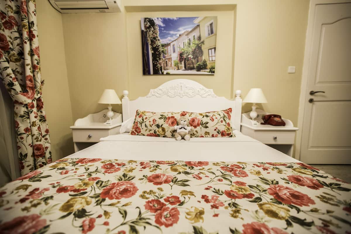 A bedroom with a floral bedspread, a white headboard, two bedside lamps, and a painting of a street scene on the wall.