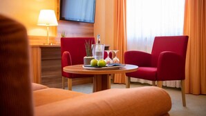 Une chambre d’hôtel avec une table ronde en bois, des chaises rouges, un téléviseur fixé au mur et une fenêtre avec des rideaux orange.