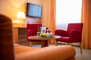 Quarto de hotel com mesa redonda de madeira, cadeiras vermelhas, televisão fixada na parede e janela com cortinas laranja.