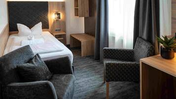 A hotel room with a bed, two armchairs, a small table, a nightstand, a wall clock, and a potted plant.