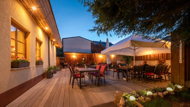 Une terrasse extérieure dotée d’un plancher en bois, de tables et de chaises, entourée d’un bâtiment rustique avec de grandes fenêtres et des plantes en pot.