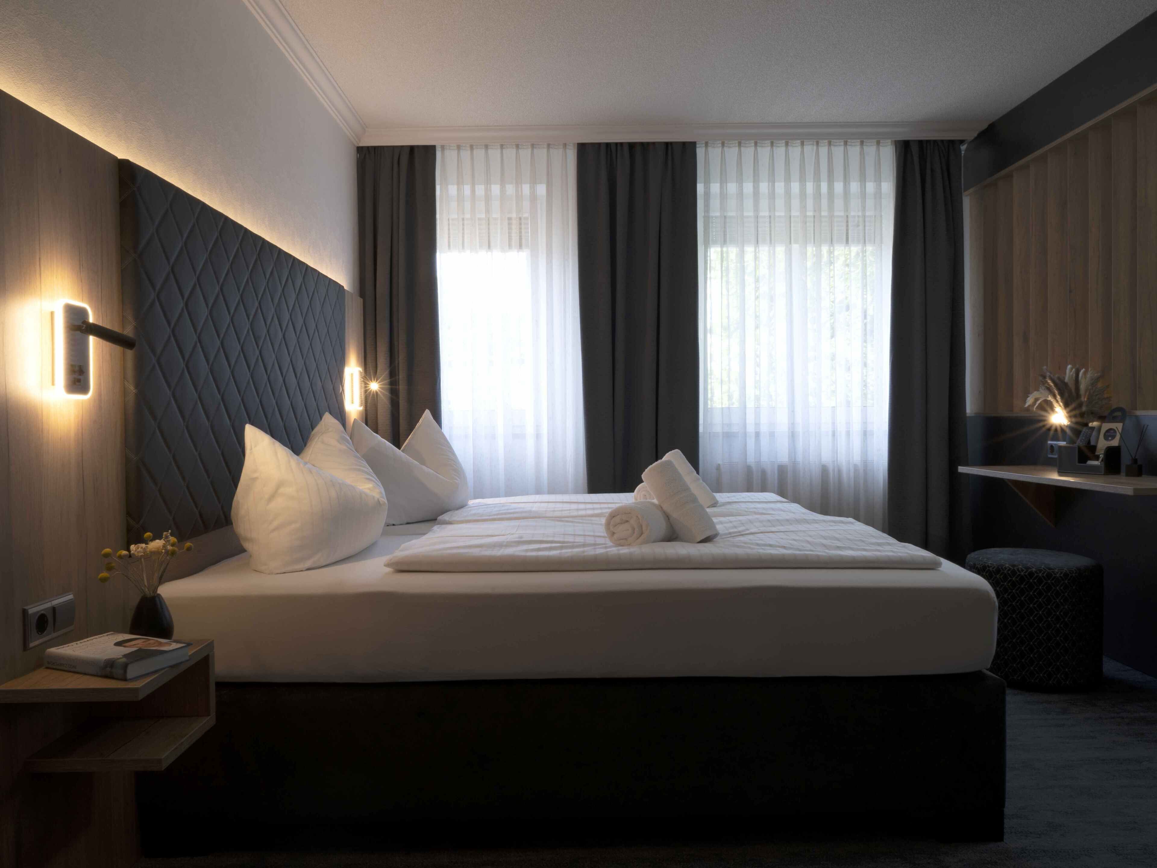 Une chambre d’hôtel moderne avec un grand lit, une tête de lit sombre, des tables de chevet et une vue sur la ville à travers de fines rideaux.