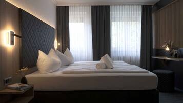A modern hotel room with a large bed, dark headboard, bedside tables, and a view of the city through sheer curtains.