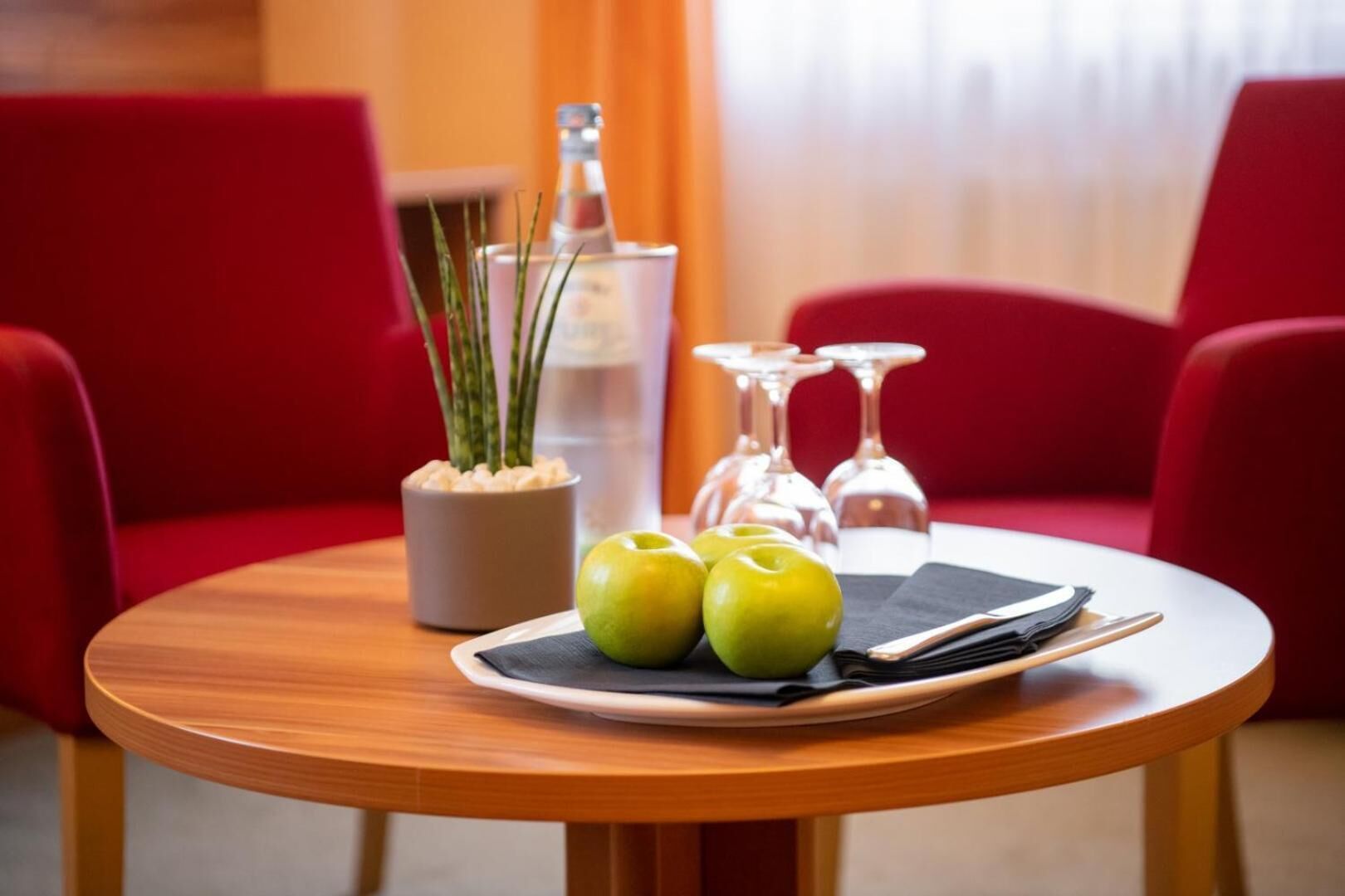 A round wooden table with a plate of green apples, a napkin, and cutlery, a glass vase with green plants, and two wine glasses.