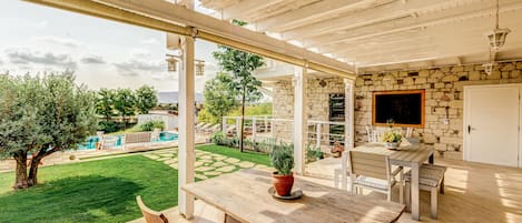 Un patio al aire libre con una mesa y sillas de madera, un muro de piedra y vistas a una piscina y jardín.