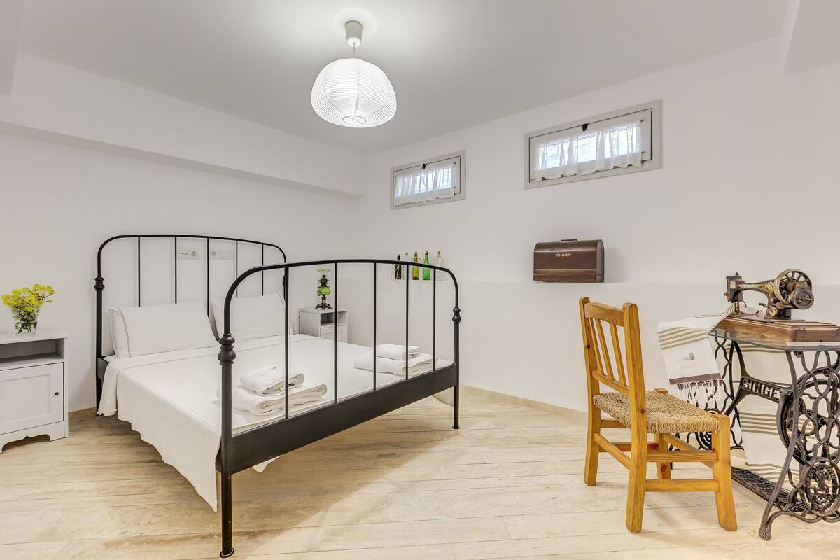 Een slaapkamer met een zwart metalen bed, witte beddengoed, een houten stoel, een vintage naaimachine en een kleine tafel met een lamp.
