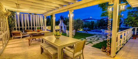 An outdoor patio with a wooden table and chairs, a white railing, and a view of a house and garden at dusk.