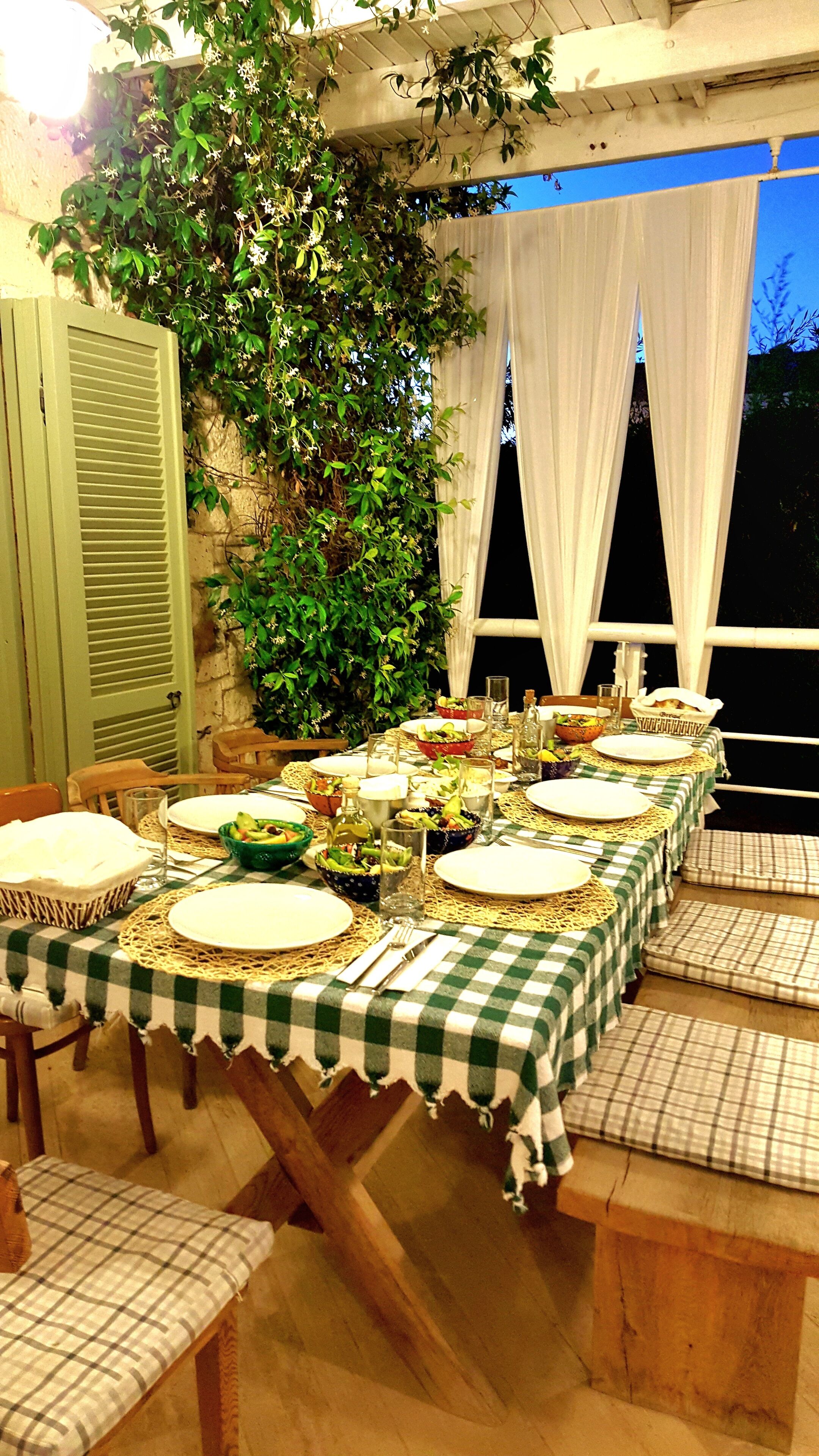 An outdoor dining area with a checkered tablecloth, wooden chairs, and a backdrop of greenery and white curtains.