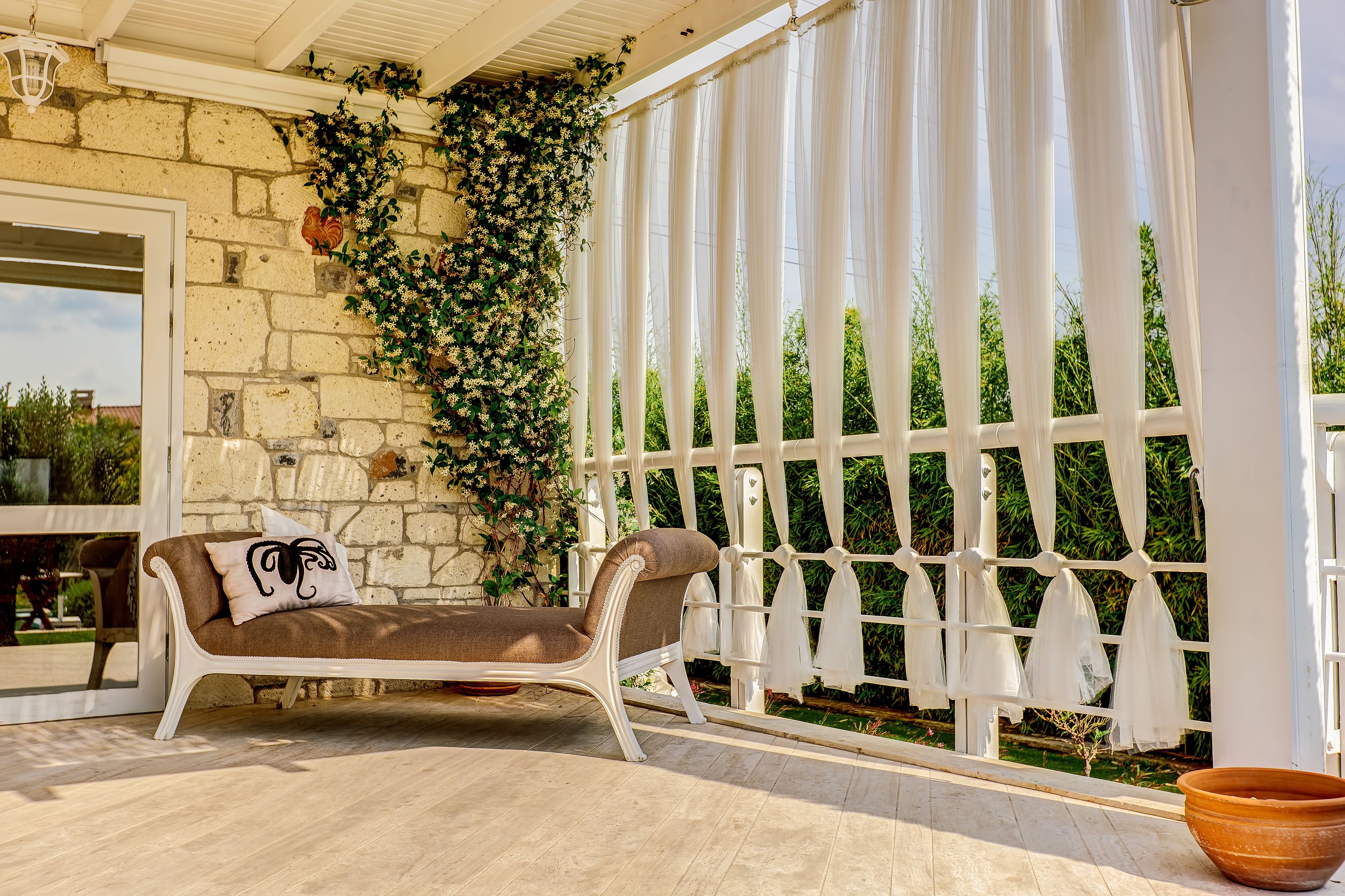 A sunlit patio with a cushioned lounge chair, a stone wall, and a white trellis with climbing plants.