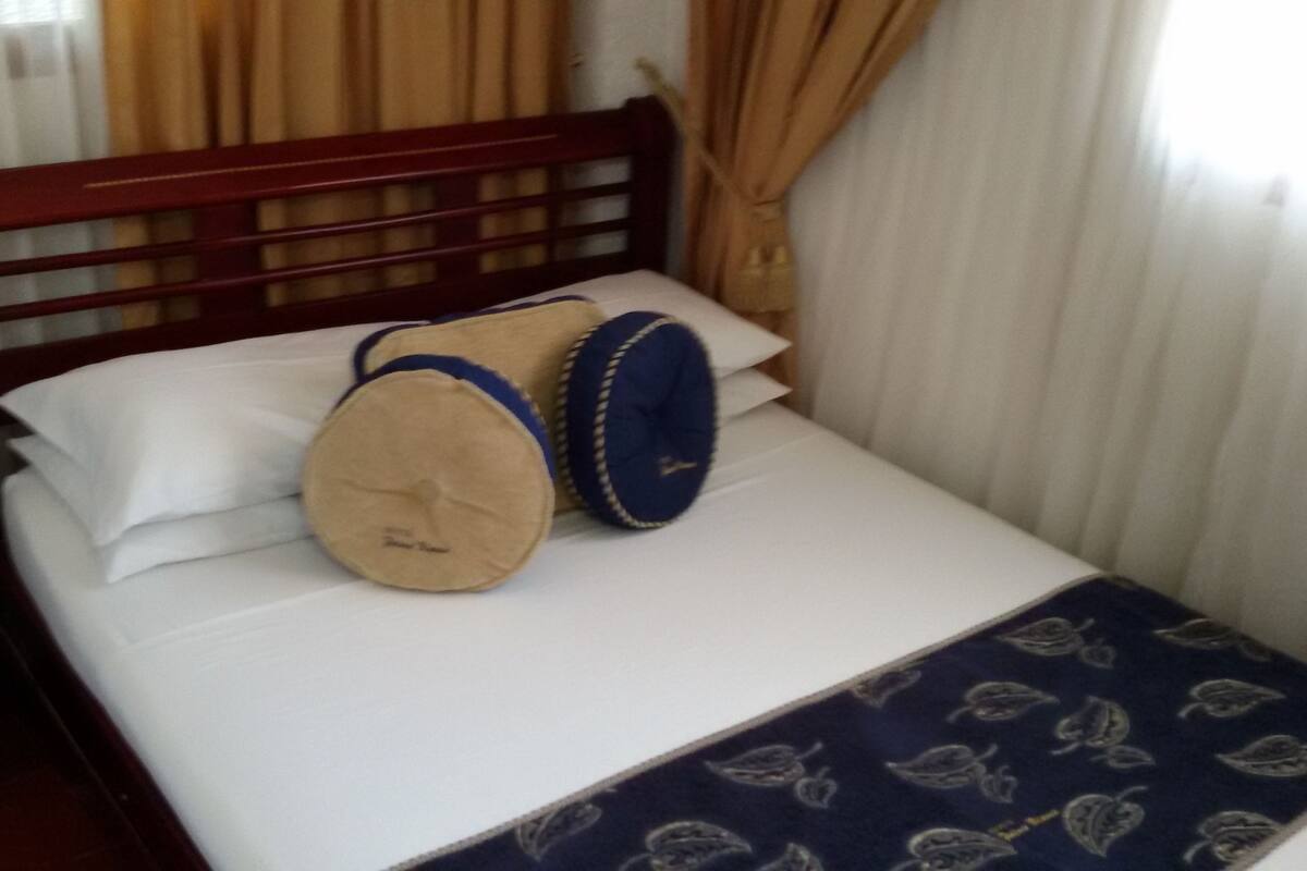 Un lit avec une tête de lit en bois, deux oreillers, un drap blanc et une couverture bleu foncé à motifs de feuilles.