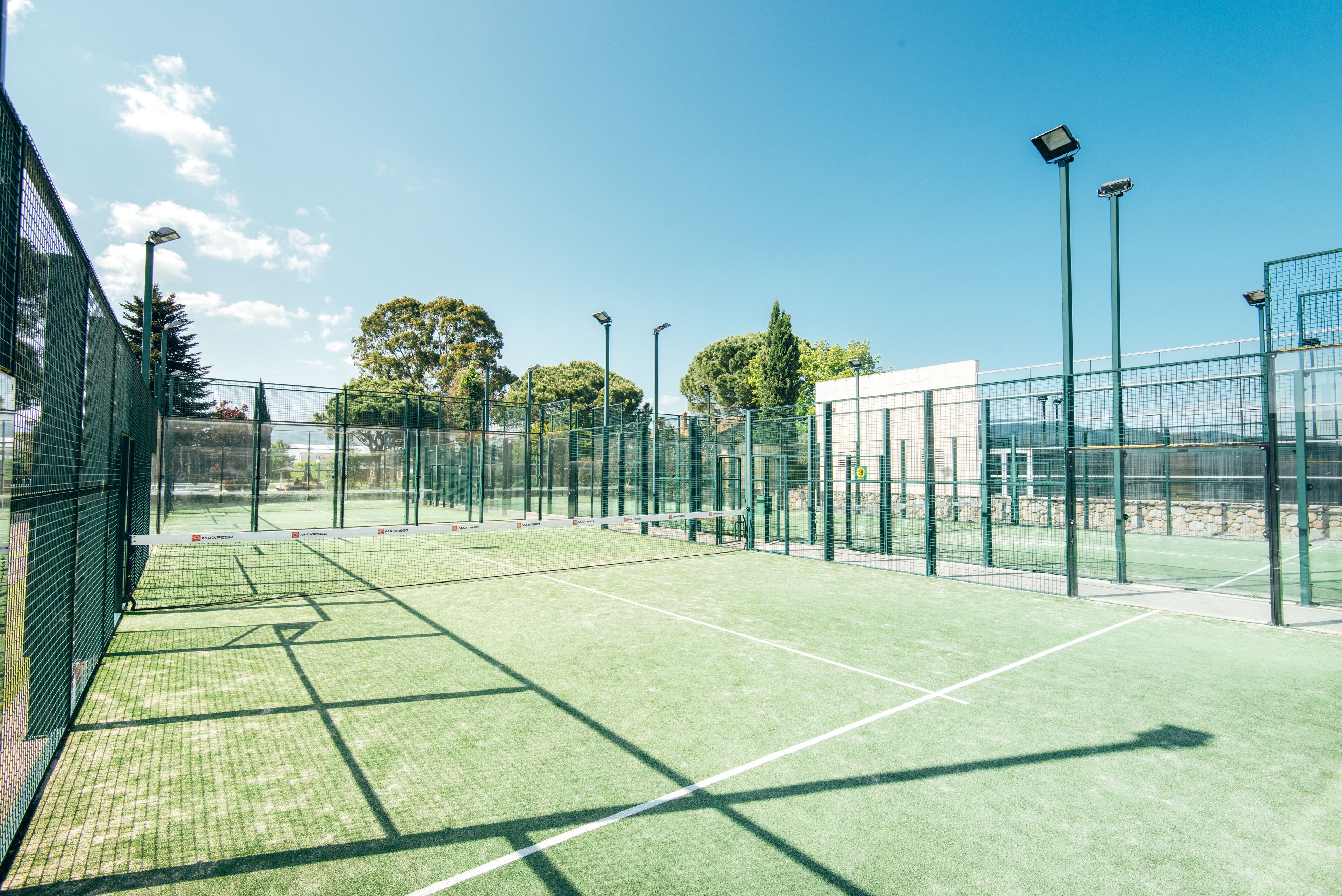 Een indoor tennisbaan met groen kunstgras, omringd door een metalen hek en voorzien van verlichting.