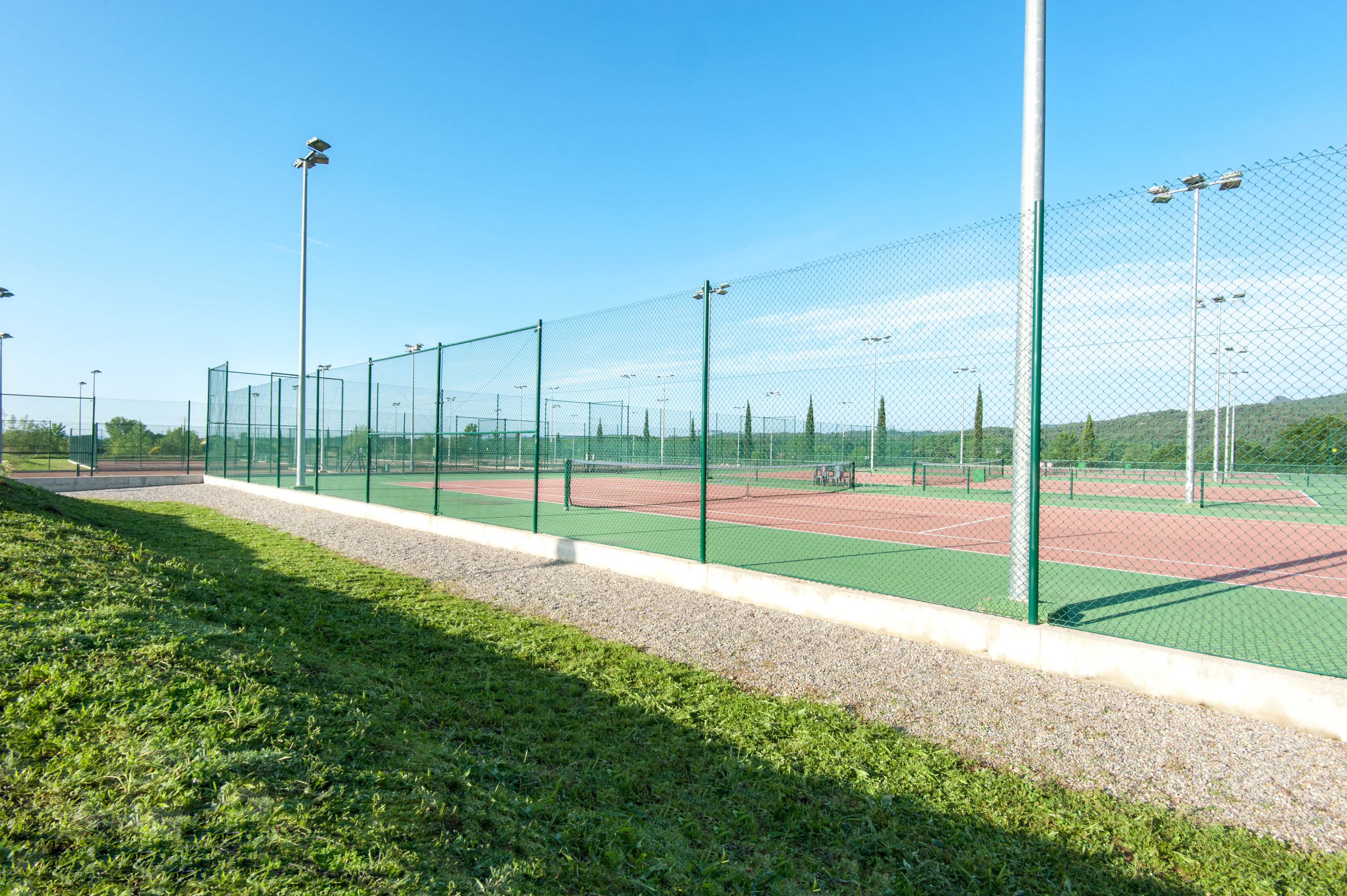 Cancha de tenis con una cerca de malla metálica, rodeada de césped y árboles.