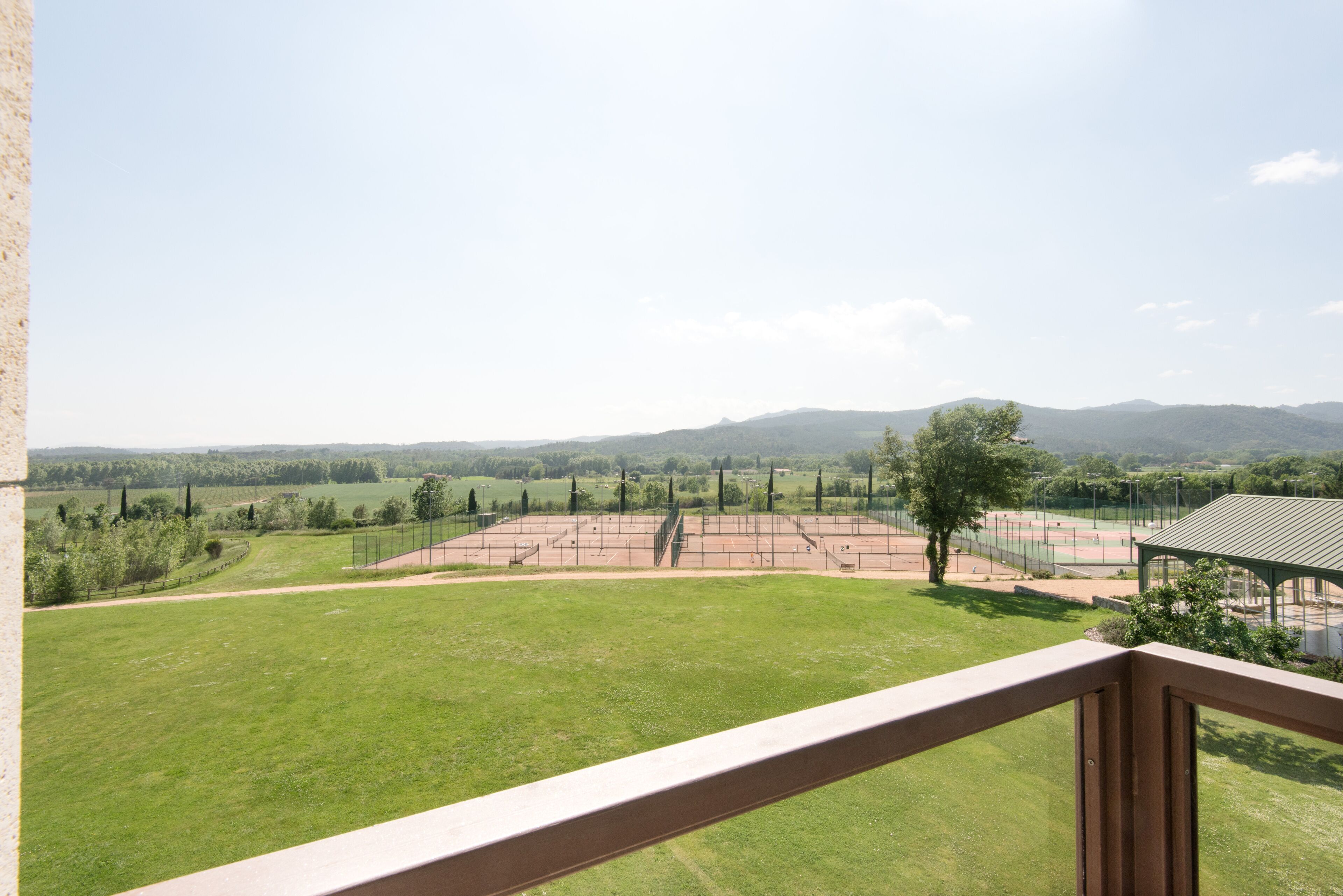 Une vue depuis un balcon donnant sur un court de tennis avec un pavillon, le tout entouré de champs verdoyants et de collines lointaines.