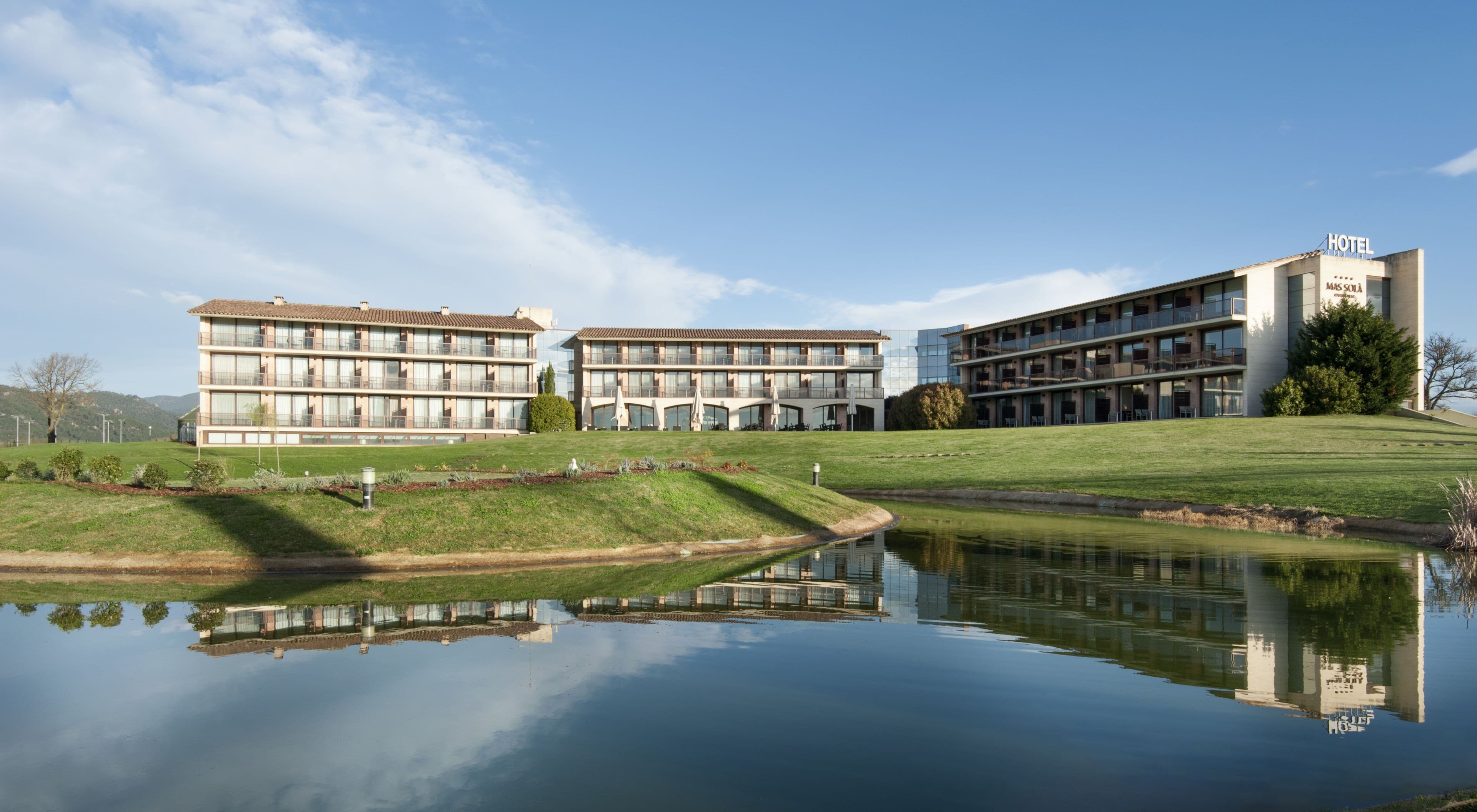 Un hôtel avec un bassin d’eau aux reflets changeants, des bâtiments modernes et un ciel dégagé.