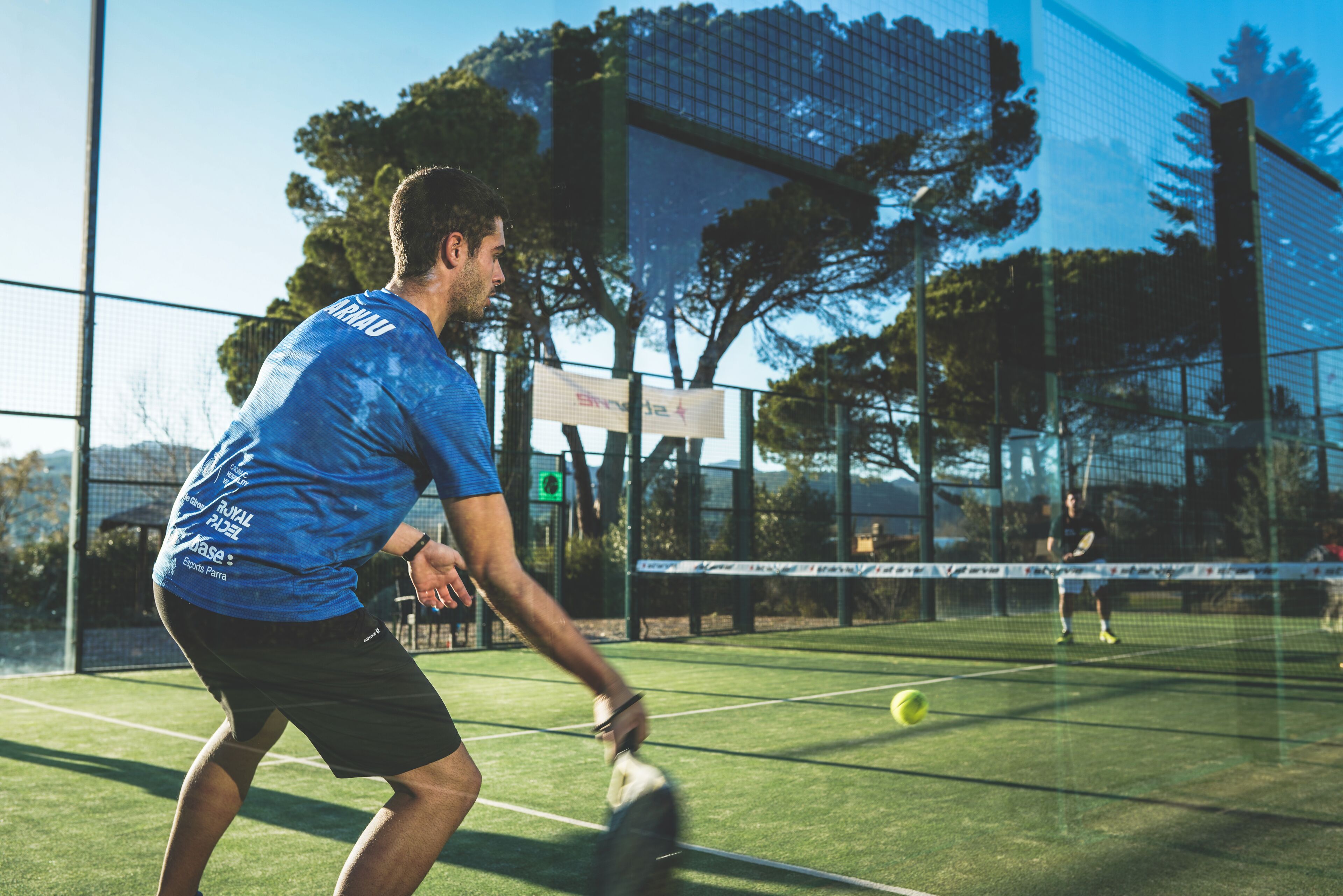A tennis court with a net, a player in a blue shirt, and another player in the background.