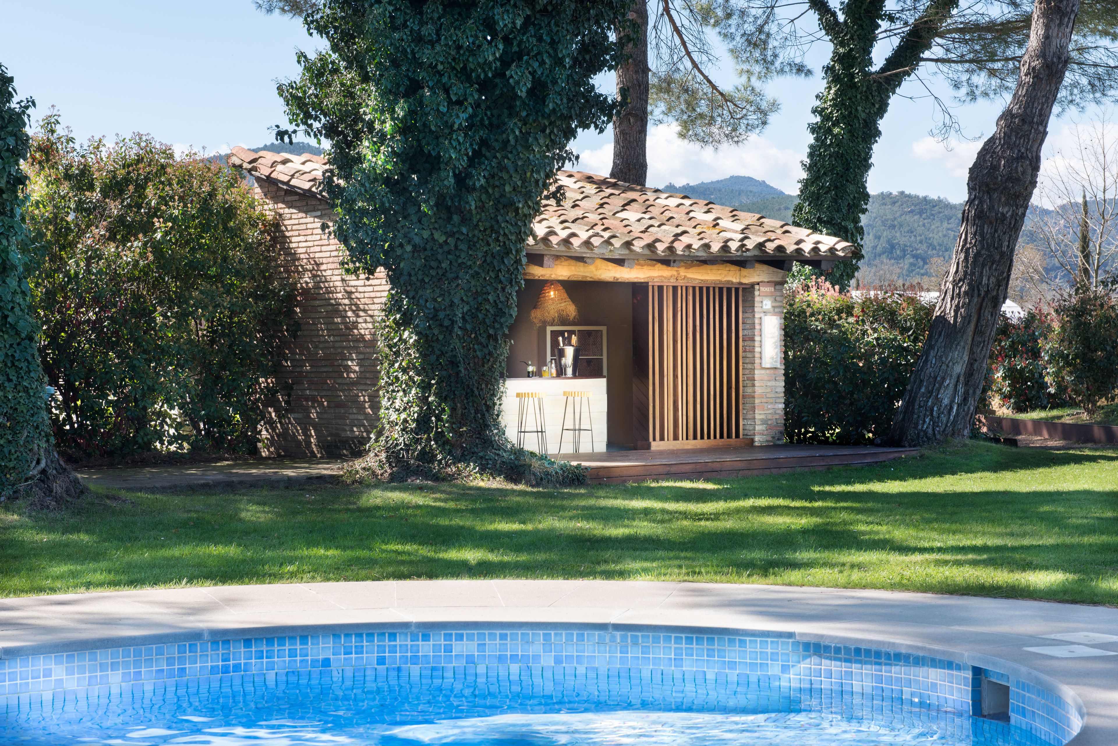 Un área de piscina con borde de azulejos, un pequeño edificio con techo de tejas y un césped.