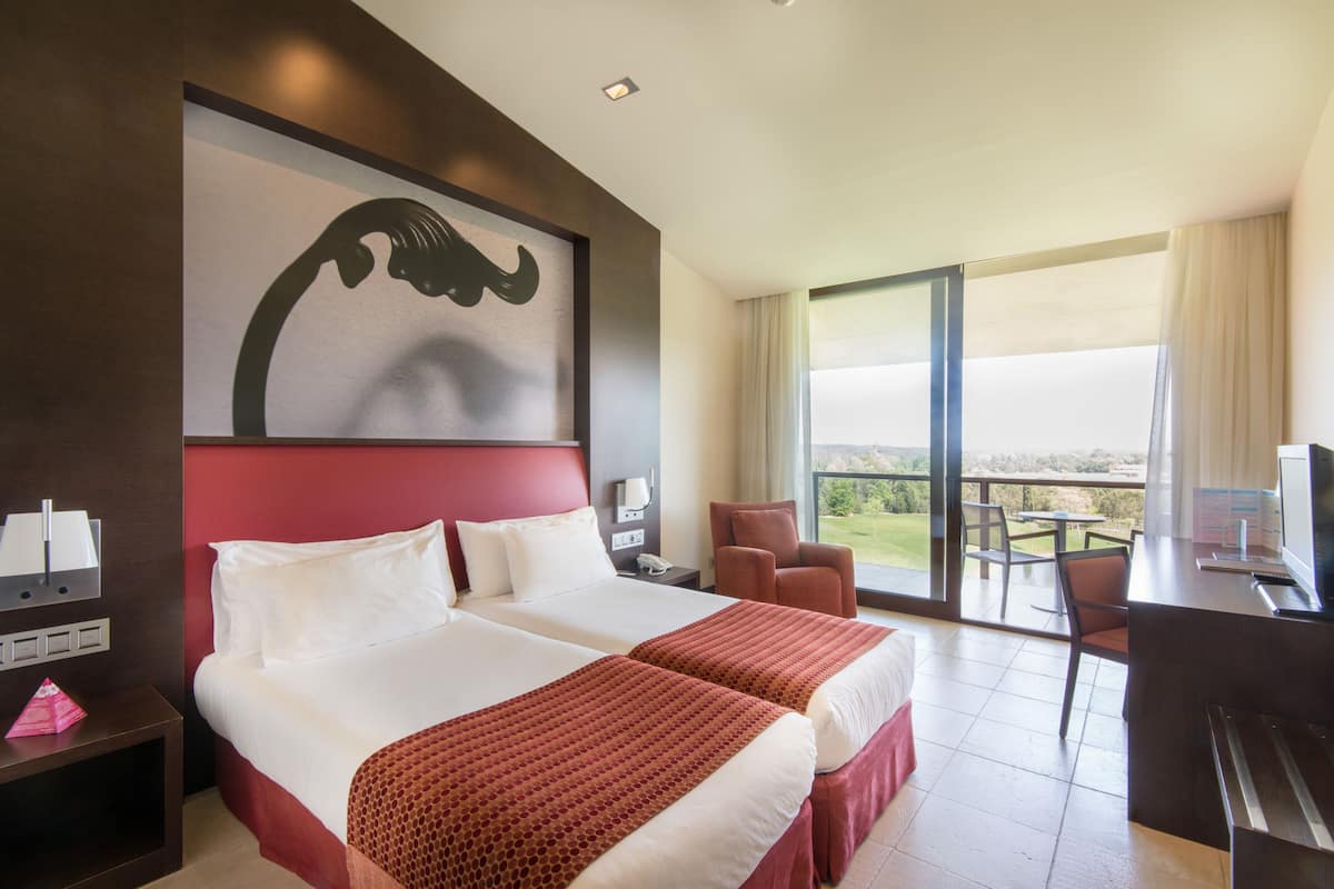 A hotel room with two beds, a large window, a balcony, and a desk with a computer.