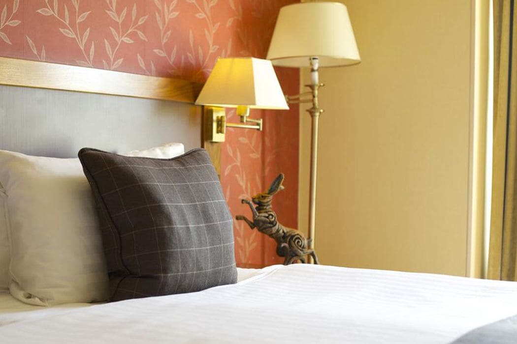 A hotel room with a bed, pillows, a lamp, and a wall-mounted light fixture.