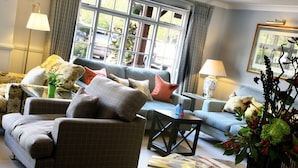 Ein gemütliches Wohnzimmer mit Sofa, Sessel und Couchtisch. Auf dem Tisch liegen Zeitungen, ein Blumenvasen und eine Lampe.