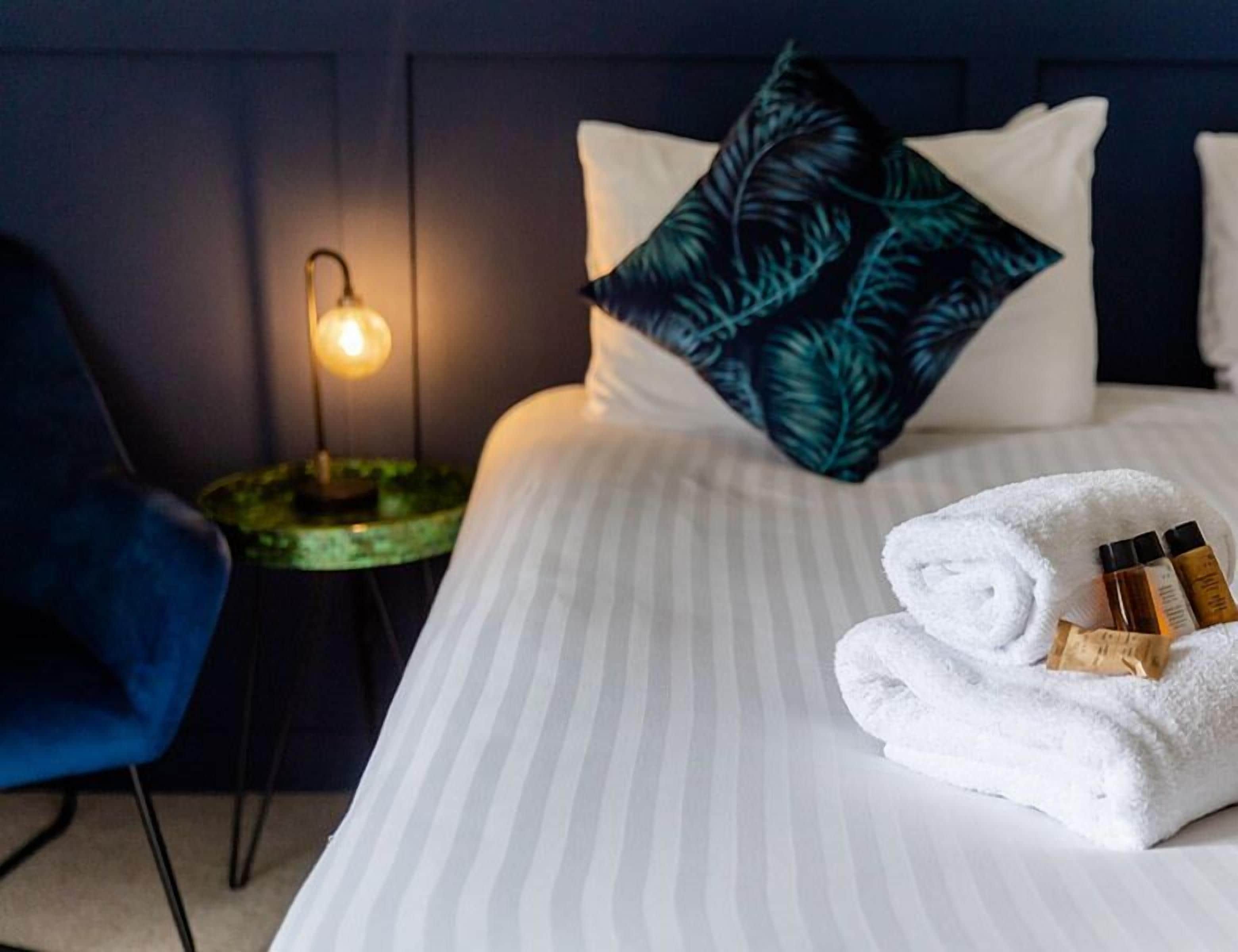 A neatly made bed with a striped bedspread, a pillow with a leaf pattern, a blue chair, and a bedside lamp.