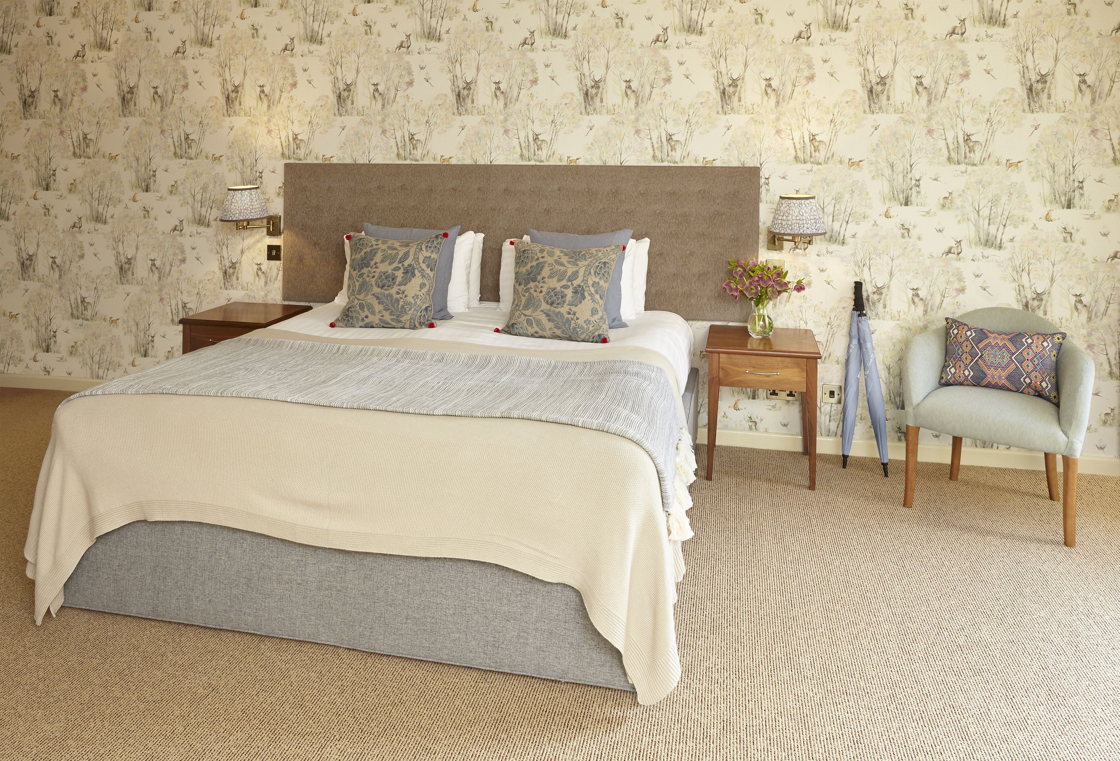 A neatly made bed with a grey headboard and patterned pillows, a small bedside table with a lamp, and a chair with a patterned cushion.