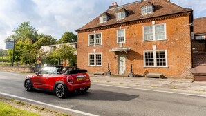 Ein roter Cabriofahrwagen vor einem Backsteinhaus mit traditioneller Gestaltung, einer sichtbaren Tür und einem kleinen Fenster im oberen Geschoss.
