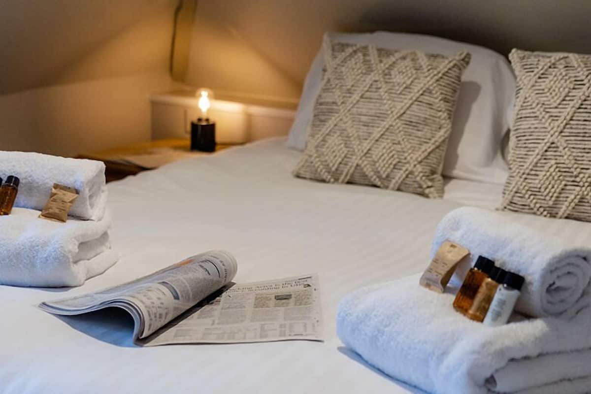 A neatly made bed with rolled towels, a newspaper, and a small bottle of lotion on the side.